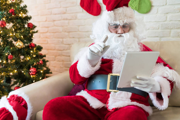 Man in Santa Claus costume using digital tablet on sofa at home - Fotografie, Obrázek