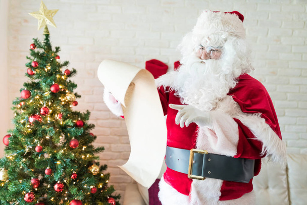 Santa Claus sprawdzanie lista chwila stojący przy Choinka w dom podczas świąteczny pora - Zdjęcie, obraz