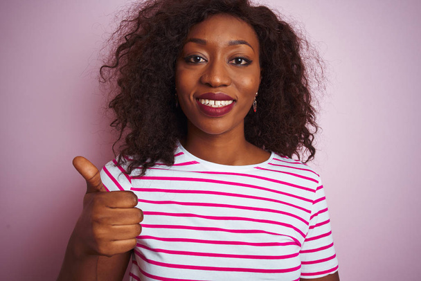 Mujer afroamericana joven con camiseta a rayas de pie sobre un fondo rosa aislado haciendo un gesto feliz con los pulgares hacia arriba con la mano. Aprobar la expresión mirando a la cámara con éxito
. - Foto, Imagen