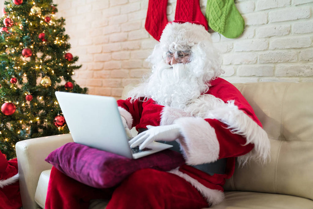 Homem vestindo traje de Papai Noel usando laptop enquanto sentado no sofá pela árvore de Natal em casa
 - Foto, Imagem