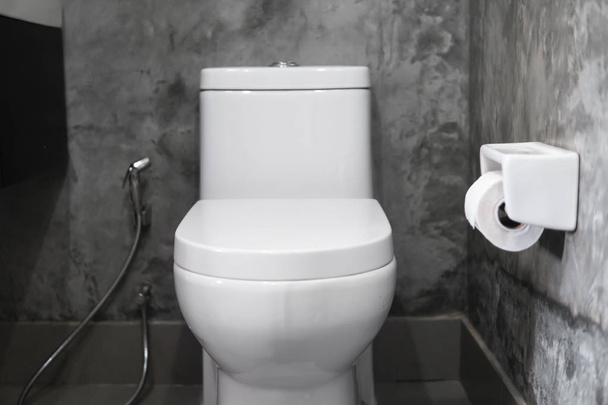 Valkoinen roikkuva wc-istuin valkoisella wc: llä kodin kylpyhuoneessa, jossa on harmaat laatat betonityylillä ja wc-paperi seinällä. Kylpyhuone ylellinen sisustus
. - Valokuva, kuva