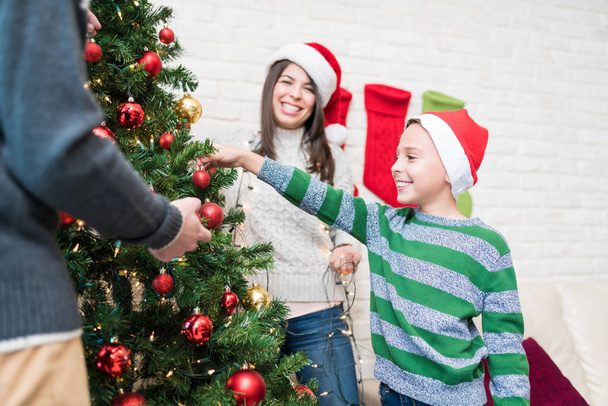 Úsměv malého kluka, který zdobí vánoční stromek s rodiči doma - Fotografie, Obrázek