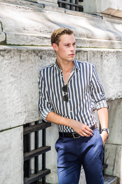 Jonge Amerikaanse man ontspannen buiten in New York City, het dragen van blanco, wit gestreept, shirt met lange mouwen, blauwe broek, zonnebril opknoping op de voorkant van shirt, staande tegen muur, frowned, verdrietig, denken - Foto, afbeelding