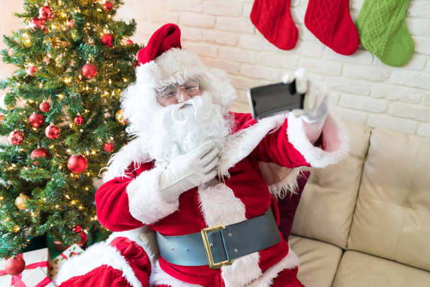 Noel Baba kostümü ve beyaz sakallı adam Noel sırasında evde akıllı telefon selfie çekerken - Fotoğraf, Görsel