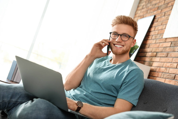 Young man talking on phone while using laptop in living room - Foto, Bild