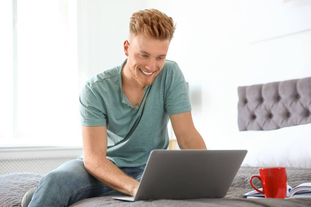 Young man using laptop while sitting on bed at home - Fotoğraf, Görsel