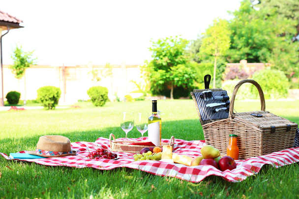 Picnic basket with products and bottle of wine on checkered blanket in garden - Фото, изображение