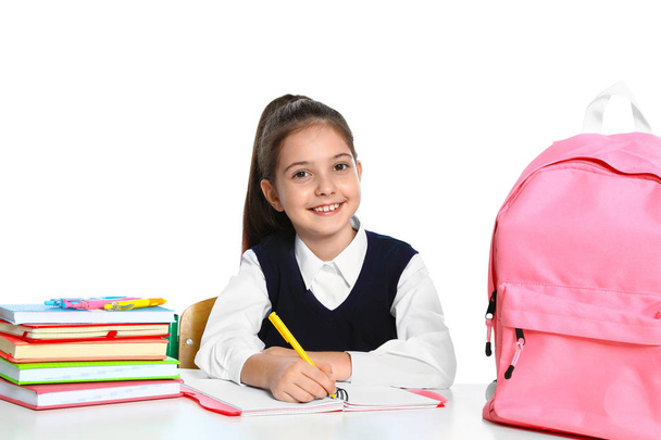 Little girl in uniform doing assignment at desk against white background. School stationery - Фото, изображение