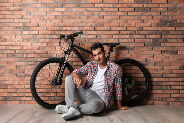 Handsome young man with modern bicycle near brick wall indoors - Фото, изображение