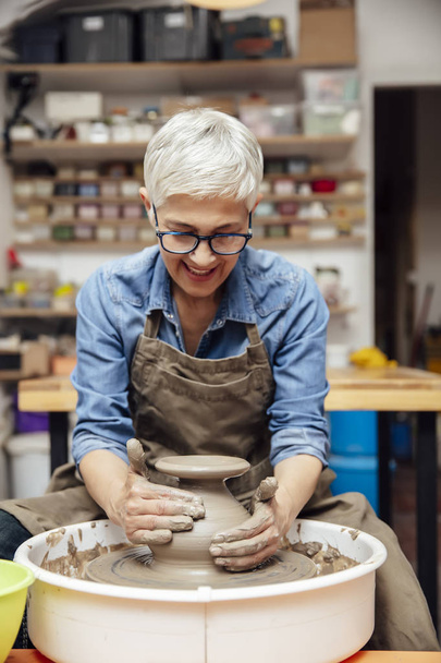Ceramista donna anziana di bell'aspetto che lavora sulla ruota della ceramica mentre è seduta nel suo laboratorio
 - Foto, immagini