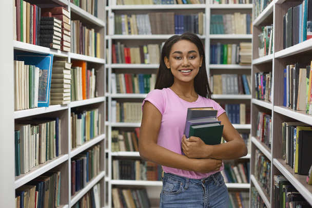 Весела чорна дівчина стоїть між книжковими полицями в бібліотеці
 - Фото, зображення