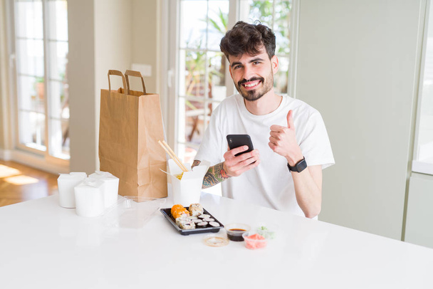 Young man eating asian sushi from home delivery and ordering food using smartphone app happy with big smile doing ok sign, thumb up with fingers, excellent sign - Photo, Image