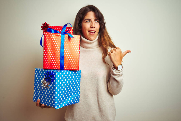 Young beautiful woman holding birthday gifts standing over isolated white background pointing and showing with thumb up to the side with happy face smiling - 写真・画像