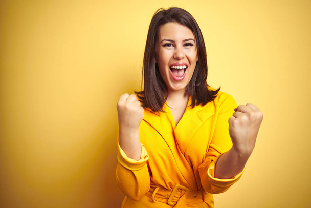 Young beautiful brunette woman wearing elegant dress over yellow isolated background celebrating surprised and amazed for success with arms raised and open eyes. Winner concept. - Photo, Image