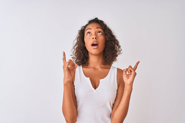 Νεαρή Βραζιλιάνα γυναίκα φορώντας casual t-shirt στέκεται πάνω από απομονωμένο λευκό φόντο έκπληκτος και έκπληκτος κοιτάζοντας προς τα πάνω και δείχνοντας με τα δάχτυλα και σήκωσε τα χέρια. - Φωτογραφία, εικόνα