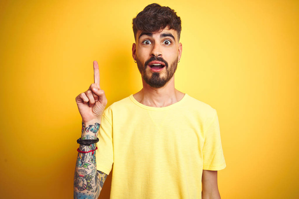 Молодой человек с татуировкой в футболке стоит на изолированном жёлтом фоне, показывая пальцем на успешную идею. Взволнован и счастлив. Номер один
. - Фото, изображение