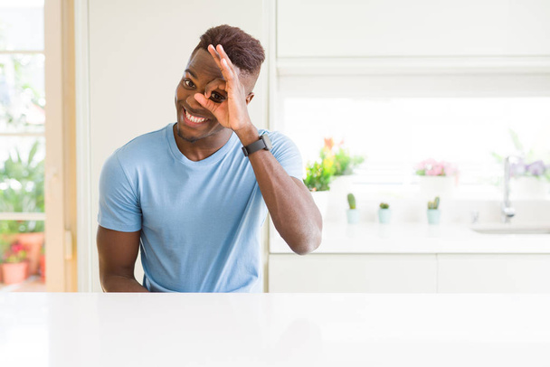 自宅でカジュアルなTシャツを着ているハンサムなアフリカ系アメリカ人男性は、手を笑顔でOKジェスチャーを行い、幸せな顔で指を通して目を見ます. - 写真・画像