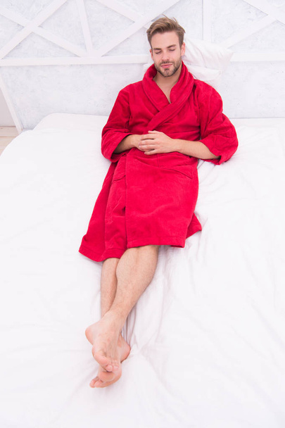 Wissen, wie man sich entspannt. Schöner Mann, der im Bett liegt, um sich nach dem Bad zu entspannen. Schläfriger Typ im roten Bademantel, der sich morgens Zeit nimmt, um sich zu entspannen. Zeit zum Entspannen - Foto, Bild