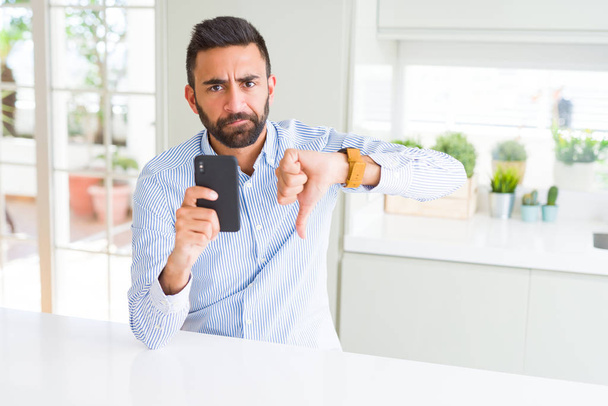 Όμορφος Ισπανόφωνος επιχειρηματίας που χρησιμοποιεί smartphone με θυμωμένο πρόσωπο, αρνητικό σημάδι που δείχνει αντιπάθεια με τους αντίχειρες κάτω, έννοια απόρριψης - Φωτογραφία, εικόνα