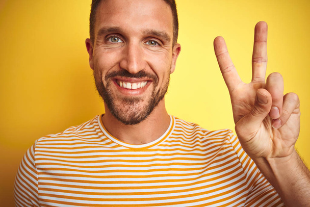 Close up of young handsome man wearing casual t-shirt over yellow isolated background smiling with happy face winking at the camera doing victory sign. Number two. - Photo, Image