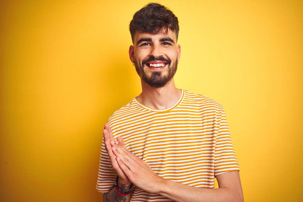 Νέος άντρας με τατουάζ φορώντας ριγέ t-shirt στέκεται πάνω από απομονωμένο κίτρινο παλαμάκια και χειροκροτώντας ευτυχισμένα και χαρούμενα, χαμογελαστά περήφανα χέρια μαζί - Φωτογραφία, εικόνα