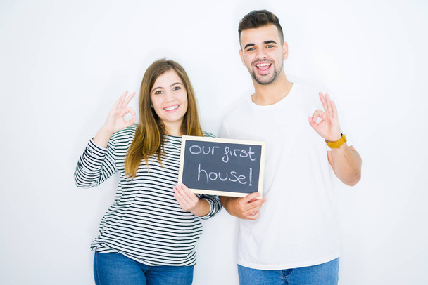 Νεαρό ζευγάρι κρατώντας μαυροπίνακα με το πρώτο κείμενο στο σπίτι μας πάνω από λευκό απομονωμένο φόντο κάνει ok υπογράψει με τα δάχτυλα, εξαιρετικό σύμβολο - Φωτογραφία, εικόνα