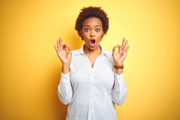 Femme d'affaires afro-américaine sur fond jaune isolé regardant surpris et choqué faisant ok symbole d'approbation avec les doigts. Expression folle
 - Photo, image
