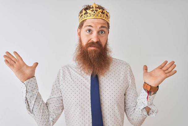 Jonge Redhead Ierse zakenman dragen kroon koning over geïsoleerde witte achtergrond clueless en verward uitdrukking met armen en handen verhoogd. Doubt concept. - Foto, afbeelding