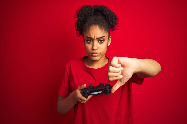 African american gamer woman playing video game using joystick over isolated red background with angry face, negative sign showing dislike with thumbs down, rejection concept - 写真・画像