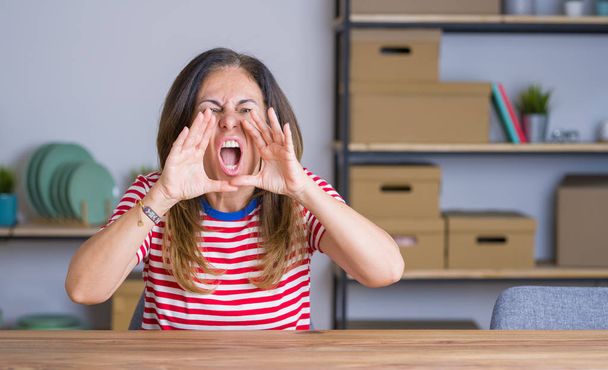 Middelbare leeftijd Senior vrouw zittend aan de tafel thuis schreeuwen boos uit luid met handen over de mond - Foto, afbeelding