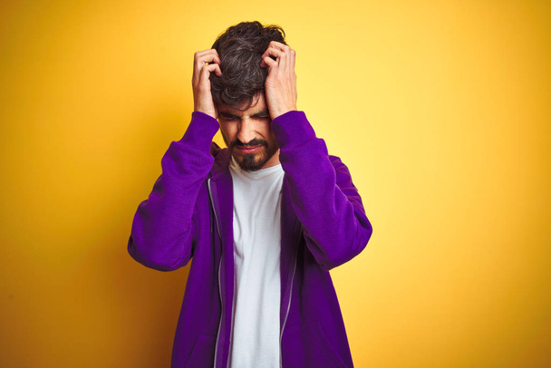 Mladý muž s tetováním na sobě nosí sportovní purpurové tričko přes izolované žluté pozadí trpící zoufalou bolestí a tlakem, protože bolest a migréna. Ruce na hlavu. - Fotografie, Obrázek