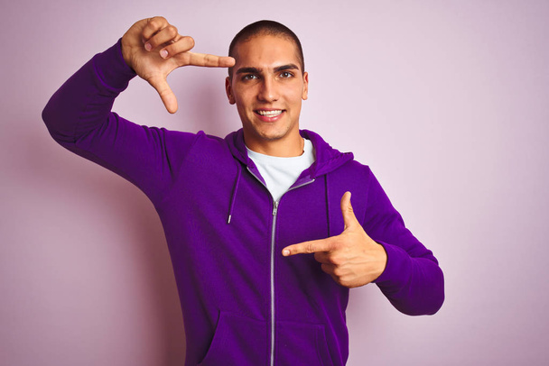 ピンクの隔絶された背景に紫色のスウェットシャツを着た若いハンサムな男が、幸せな顔をした手と指でフレームを作っている。創造性と写真の概念. - 写真・画像