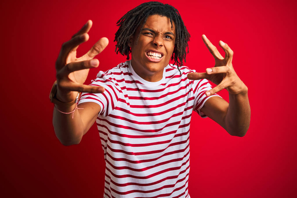 Mladý Afro muž s dredy v proužkované košili stojící nad izolovaným červeným pozadím křičící vztek, ruce snaže se uškrtit a křičet - Fotografie, Obrázek