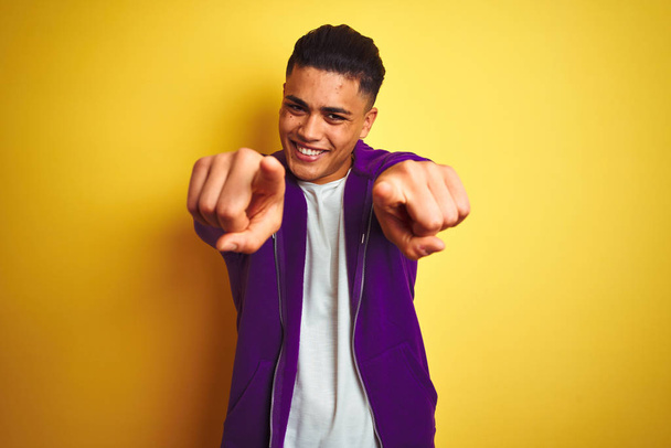 Молодой бразилец в фиолетовой толстовке стоит на изолированном желтом фоне, указывая на тебя и камеру пальцами, улыбаясь позитивно и весело
 - Фото, изображение