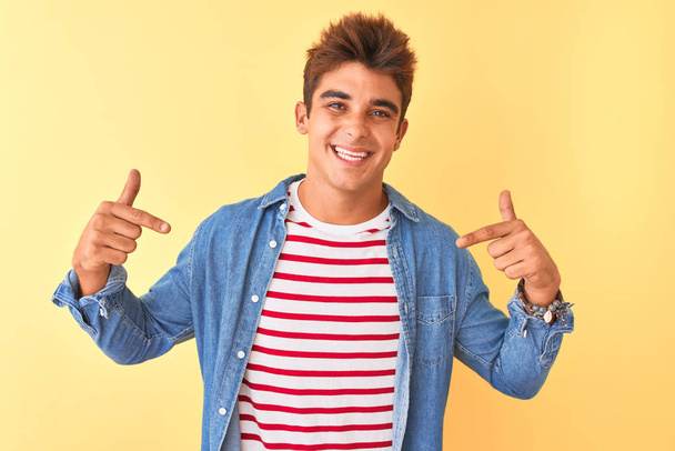 Young handsome man wearing striped t-shirt and denim shirt over isolated yellow background looking confident with smile on face, pointing oneself with fingers proud and happy. - Photo, Image