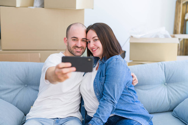 新しい家のソファに座っている若いカップルは、新しいアパートに引っ越すために幸せに笑顔のスマートフォンを使用して自分撮りを取ります - 写真・画像