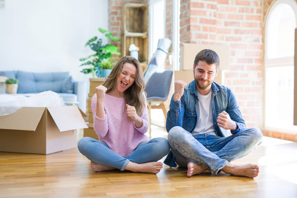Fiatal szép pár költözött egy új ház ül a padlón nagyon boldog és izgatott csinál győztes gesztus a fegyvert emelt, mosolyogva és sikítva a sikerhez. Ünneplés koncepció. - Fotó, kép
