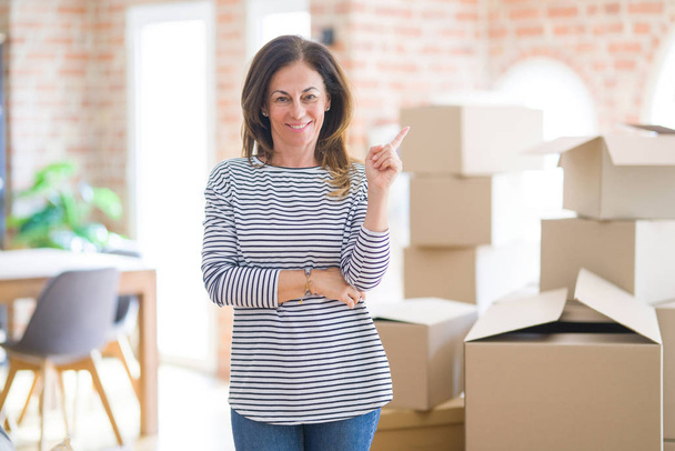 Женщина средних лет переезжает в новый дом вокруг картонных коробок с широкой улыбкой на лице, указывая рукой и пальцем в сторону, глядя на камеру
. - Фото, изображение