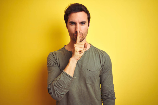Young handsome man wearing casual t-shirt standing over isolated yellow background asking to be quiet with finger on lips. Silence and secret concept. - Photo, Image