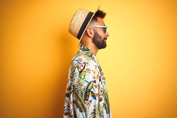 Hombre indio de vacaciones con gafas de sol de sombrero de camisa floral sobre fondo amarillo aislado mirando a un lado, pose de perfil relajado con cara natural con sonrisa confiada
. - Foto, Imagen