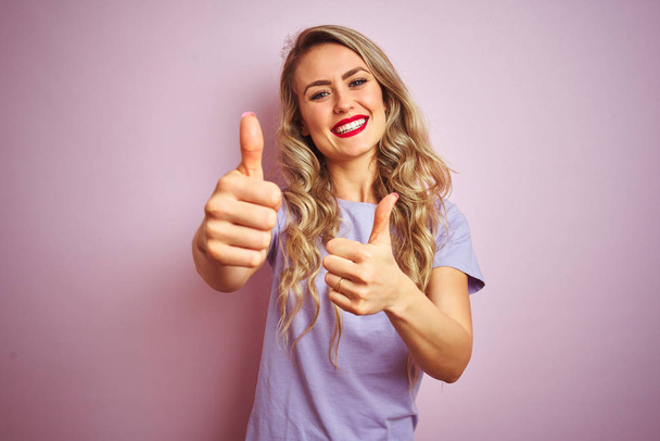 junge schöne Frau in lila T-Shirt vor rosa isoliertem Hintergrund stehend, zustimmend positive Geste mit der Hand machend, Daumen hoch lächelnd und glücklich über den Erfolg. Siegergeste. - Foto, Bild