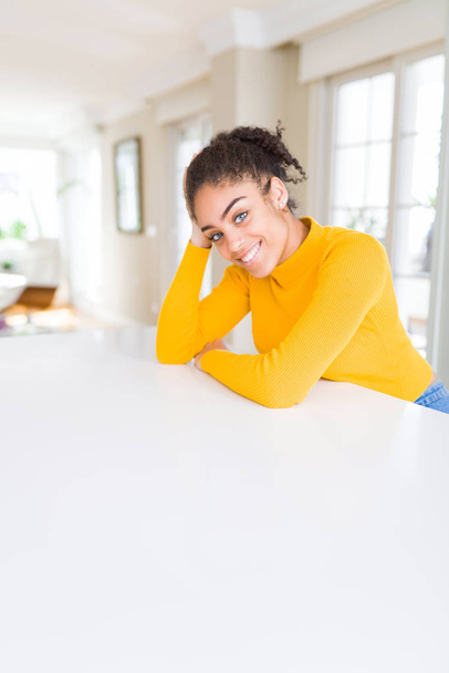 Mooie jonge Afro-Amerikaanse vrouw glimlachend vrolijk leunend op de tafel comfortabel - Foto, afbeelding