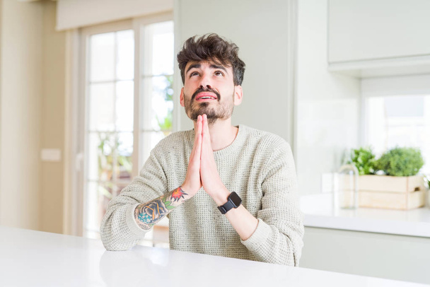 Young man wearing casual sweater sitting on white table begging and praying with hands together with hope expression on face very emotional and worried. Asking for forgiveness. Religion concept. - Photo, Image