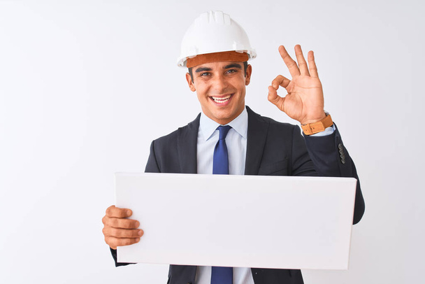 młody przystojny architekt człowiek nosić kask trzymając sztandar na odizolowanym białym tle robi ok znak palcami, doskonały symbol - Zdjęcie, obraz