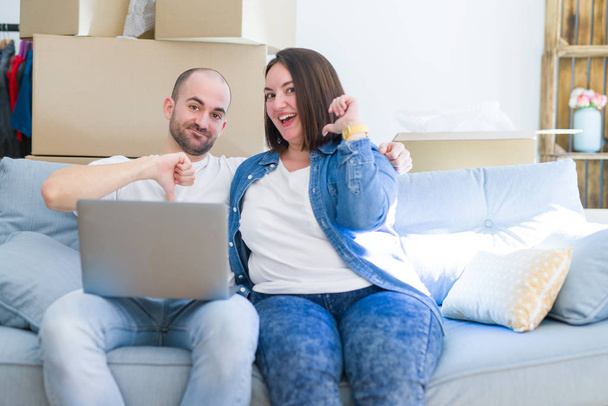 Young couple sitting on the sofa arround cardboard boxes moving to a new house using laptop with surprise face pointing finger to himself - Foto, Imagem