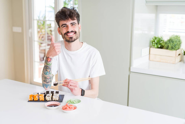 junger Mann isst asiatisches Sushi von zu Hause Lieferung glücklich mit großem Lächeln tun ok Zeichen, Daumen nach oben mit den Fingern, hervorragende Zeichen - Foto, Bild