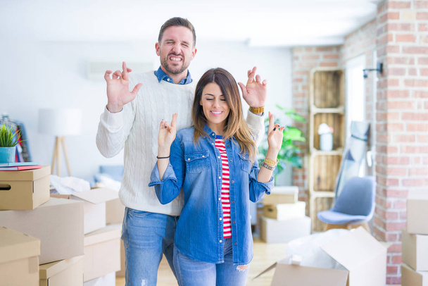 Young beautiful couple standing at new home around cardboard boxes gesturing finger crossed smiling with hope and eyes closed. Luck and superstitious concept. - Zdjęcie, obraz