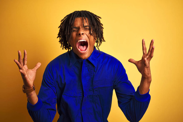 Afro worker man with dreadlocks wearing mechanic uniform over isolated yellow background celebrating mad and crazy for success with arms raised and closed eyes screaming excited. Winner concept - Valokuva, kuva