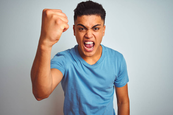 Fiatal brazil férfi visel kék póló állt át elszigetelt fehér háttér dühös és őrült emelése ököl frusztrált és dühös, miközben kiabálva a düh. Düh és agresszív koncepció. - Fotó, kép