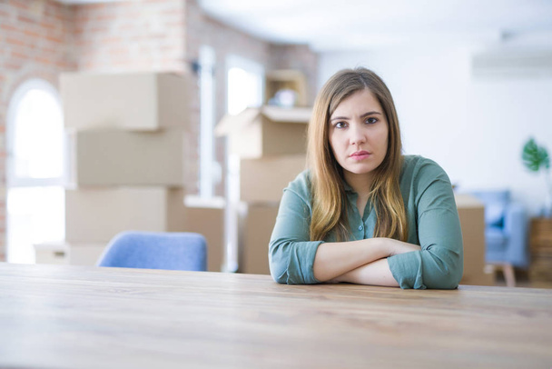 Young woman sitting on the table with cardboard boxes behind her moving to new home skeptic and nervous, disapproving expression on face with crossed arms. Negative person. - Foto, Imagen